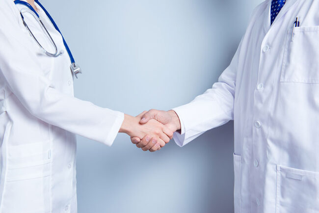 Deal! Concept of collaboration in medicine. Close up photo of two doctors’ handshake on gray background