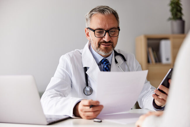 A senior doctor is reading a patient’s blood results, sitting at the office.